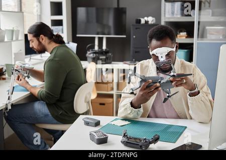 Porträt von zwei Leuten, die in einer technischen Werkstatt arbeiten, mit Fokus auf Schwarzer Mann, der Drohne inspiziert, Kopierraum Stockfoto