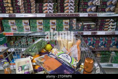 London, Großbritannien. Dezember 2023. Ein Einkaufswagen mit Weihnachtsessen in einem Sainsbury's Supermarkt in Balham. Auf den Regalen hinter dem Wagen befinden sich Hackkuchen und Weihnachtskuchen. Quelle: Anna Watson/Alamy Live News Stockfoto