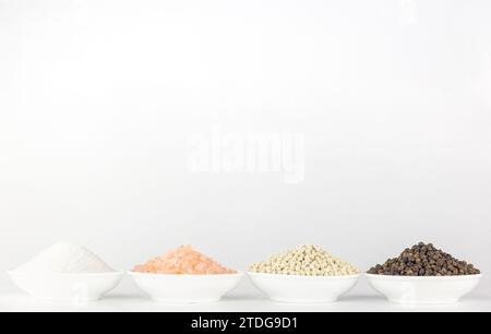 Weißes Meersalz, rosafarbenes Salz, weißer Pfeffer und schwarzer Pfeffer in separaten weißen Schüsseln auf weißem Hintergrund. Stockfoto