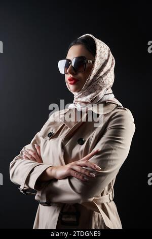 Modisches weibliches Modell in trendigem Mantel und Sonnenbrille, das im Studio wegblickt. Seitenansicht einer schweren Frau im Kopftuch stehend mit gekreuzten Armen in Dunkelheit, isoliert auf Schwarz. Modekonzept. Stockfoto