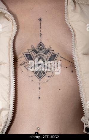 Ein kompliziertes, symmetrisches, lotosähnliches Tattoo ziert die obere Brust, eingerahmt von einer offenen strukturierten Jacke Stockfoto