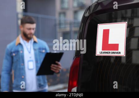 Kursleiter mit Klemmbrett in der Nähe des Autos im Freien, selektiver Fokus auf L-Platte. Fahrschule Stockfoto