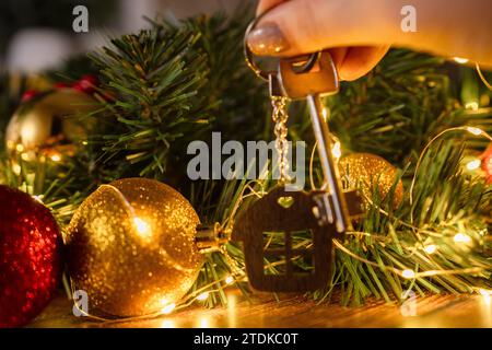 Geschenk Schlüsselhaus mit Schlüsselanhänger in der Hand Hintergrund des Weihnachtsbaums. Bauen, entwerfen, Projekt, Umzug in ein neues Haus, Hypothek, Miete und Kauf von Immobilien Stockfoto