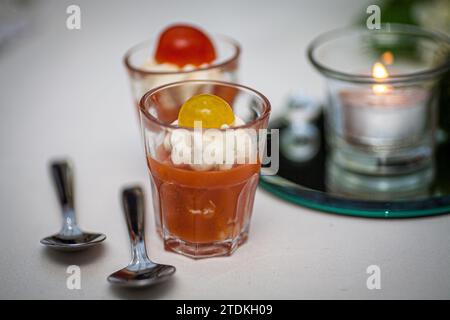 Dieses Bild zeigt eine elegante Vorspeise mit einem Glas cremiger Tomatensuppe, garniert mit einem Klecks reichhaltiger Sahne und einer Kirschtomatengarnitur. Die Suppe wird in einem klaren Glas serviert, sodass die Schichten des Gerichts visuell wahrgenommen werden können. Im weichen Hintergrund sorgt eine Kerze in einem Glashalter für ein warmes, einladendes Leuchten. Begleitet von zwei kleinen Löffeln bietet dieses Gericht einen Geschmack von raffiniertem Geschmack in einer intimen Atmosphäre. Gourmet-Vorspeise: Tomatensuppe und Creme im Glas. Hochwertige Fotos Stockfoto