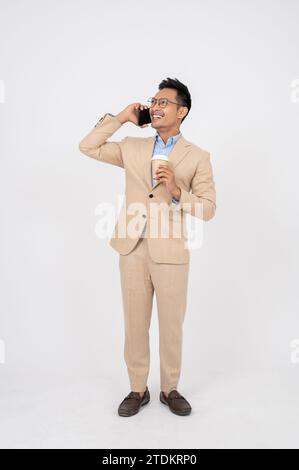 Ein gutaussehender und professioneller asiatischer Geschäftsmann hält eine Kaffeetasse in der Hand und spricht mit jemandem am Telefon, der auf einem isolierten weißen Atelierrücken steht Stockfoto