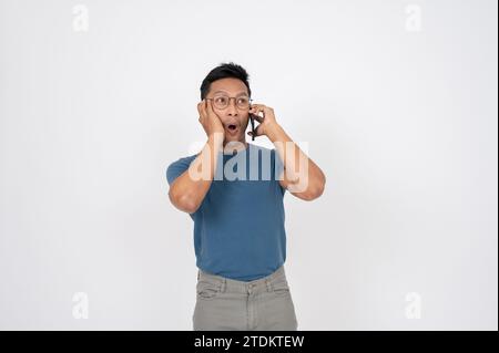 Ein überraschter, schockierter asiatischer Mann in lässiger Kleidung spricht am Telefon mit jemandem, der auf einem isolierten weißen Studio-Hintergrund steht. Unglaublich n Stockfoto