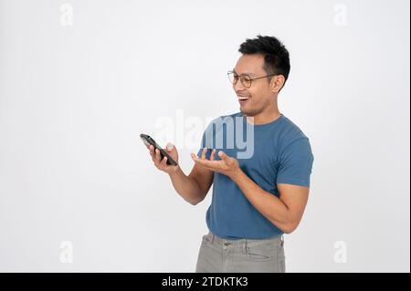 Ein positiver, glücklicher asiatischer Mann überrascht mit Nachrichten auf seinem Smartphone, das sein Telefon benutzt, und steht auf einem isolierten weißen Studio-Hintergrund. Stockfoto