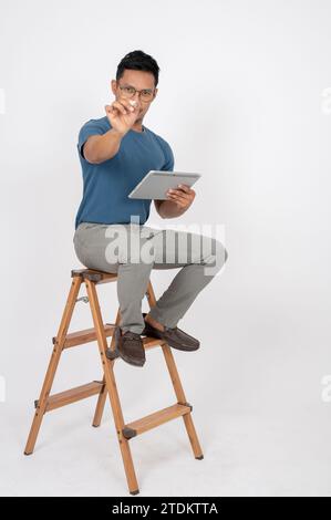 Ein asiatischer Mann in lässiger Kleidung zeigt mit seinem Stift auf die Kamera, hält sein digitales Tablet und sitzt auf einer Holzleiter auf einem isolierten weißen Hengst Stockfoto