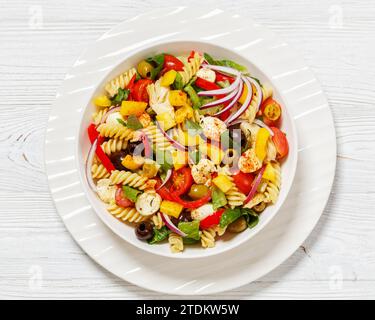 Fusilli-Salat mit Mini-Mozzarella, Tomaten, Römersalat, Oliven, Zwiebeln, Paprika und Gewürze in weißer Schüssel auf weißem Holztisch, horizonta Stockfoto