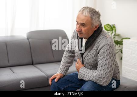 Besorgter älterer Mann, der sich schlecht fühlt, verärgert alter Großvater mittleren Alters, der die Brust berührt, plötzliche Schmerzen Sodbrennen, Herzinfarkt haben, auf dem Sofa sitzen Stockfoto