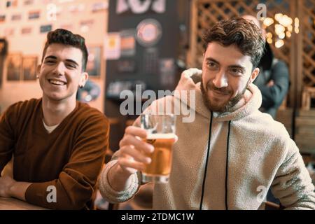 Zwei fröhliche junge Männer, die in einem gemütlichen Pub mit Biergläsern tosten und einen glücklichen Moment teilen. Stockfoto