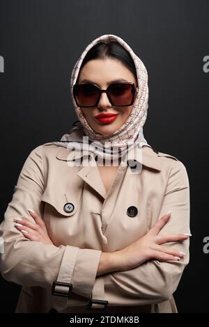 Modisches weibliches Modell in beigefarbenem Trenchcoat und Kopftuch lächelnd vor der Kamera. Porträt einer attraktiven Frau mit Sonnenbrille, die mit gekreuztem Arm posiert, vor schwarzem Hintergrund. Modekonzept. Stockfoto