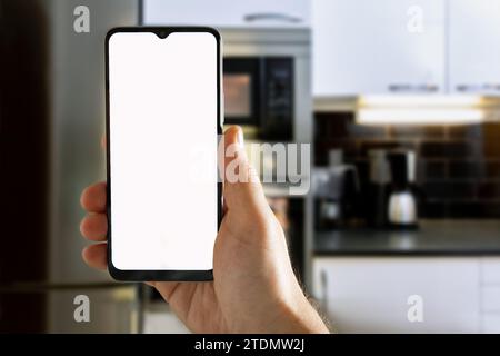 Nahaufnahme der Hand eines Mannes, der das Mobile hält, mit weißem Bildschirm für Smart Home Control Anwendungssymbole mit Küchenhintergrund Stockfoto
