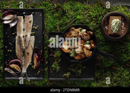 Heringsfilet mit Kapern, Salzflocken, roten Zwiebeln und Pfeffer, Waldpilzen und Brotscheibe mit Kräuterbutter serviert auf schwarzem Tablett auf grünem Gemüse Stockfoto