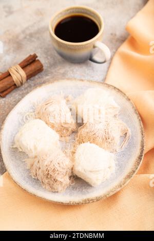 Traditionelle arabische Süßigkeiten pishmanie und eine Tasse Kaffee auf braunem Betonhintergrund und orangefarbenem Leinentuch. Seitenansicht, Nahaufnahme, selektiver Fokus. Stockfoto