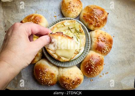 Frisch gebackene Hefeteig-Brötchen und geschmolzener Brie-Käse auf Backform, ein Stück in Menschenhand Stockfoto