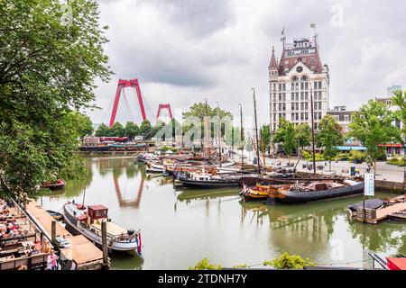 Der alte Hafen von Rotterdam mit Flussbooten, überblickt vom historischen Gebäude Witte Huis und den roten Pylonen der Willemsbrug, unter einem stürmischen Himmel. Stockfoto