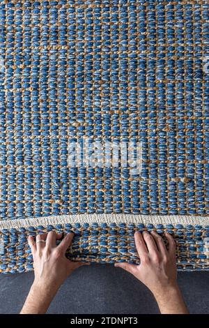 Mann, der Hand gewebte geometrische Denim-Teppich auf einem Holzboden zu Hause rollte. Stockfoto