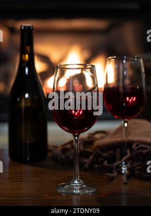 Zwei Gläser Rotwein mit Decke und Flasche auf Holztisch. Brennender Kamin im Erdgeschoss Stockfoto