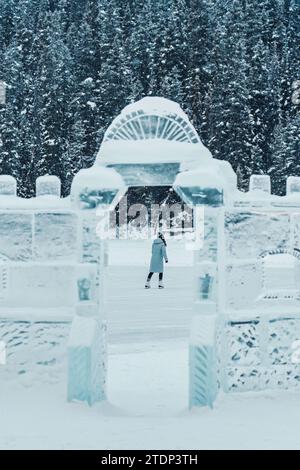 Junge Touristikerin mit Mütze im Wintermantel, die auf dem gefrorenen See Schlittschuhlaufen spielt, und Eiswand im Winterwunderland Stockfoto