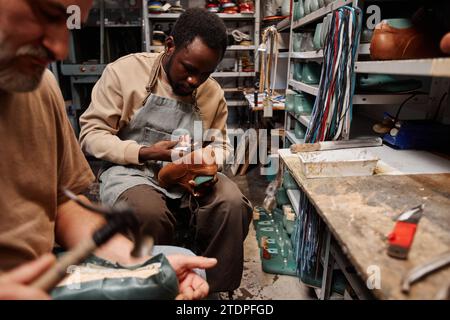 Junger afroamerikanischer Mann in Schürze, der den oberen Teil der Schuhe mit der Sohle zusammensetzt, während er über neue Schuhe oder Stiefel arbeitet Stockfoto