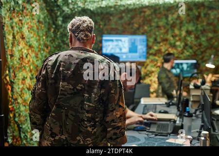 Rückansicht einer jungen Offizierin in Tarnuniform, die vor der Kamera im Überwachungsraum mit männlichen Kollegen steht Stockfoto