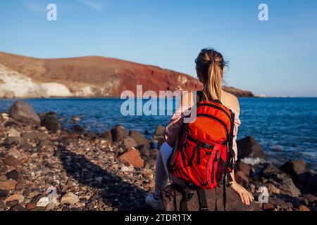 Reisende Frau mit Rucksack, die sich am Roten Strand auf Santorin, Griechenland, entspannt und die Meer- und Berglandschaft genießt. Reisen und Urlaub Stockfoto