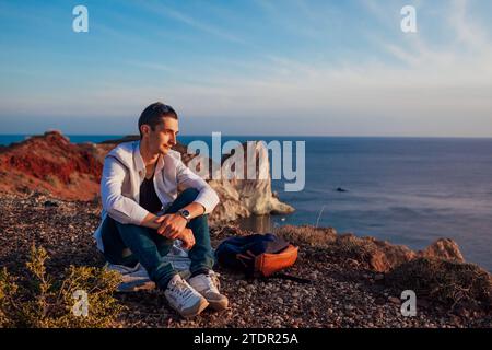 Touristen-Wanderer, die sich auf einem Berg auf dem Meer auf der Insel Santorin ausruhen und die Landschaft genießen. Glücklicher Mann bewundert den Sonnenuntergang am Meer Stockfoto