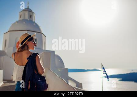 Touristenfrau, die die Caldera-Meereslandschaft in Fira, Santorini-Insel, bei einem Spaziergang durch die traditionelle weiße Kirche genießt. Griechische Architektur und Flagge Stockfoto