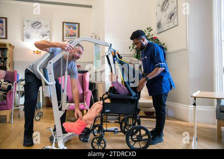 Krankenschwester Evans Sorbeng (aus Ghana) hilft dem Pfleger Jim Clarke, die Notdienerin Angela Morgan (76) in einen Stuhl im Tageszimmer des St. Cecilias Nursing Home Scarborough St. Cecilias Nursing Home Scarborough zu heben. Stockfoto