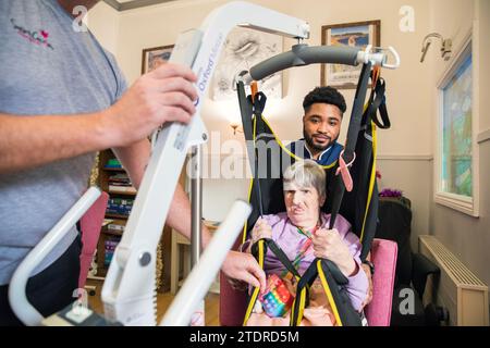 Krankenschwester Evans Sorbeng (aus Ghana) hilft dem Pfleger Jim Clarke, die Notdienerin Angela Morgan (76) in einen Stuhl im Tageszimmer des St. Cecilias Nursing Home Scarborough St. Cecilias Nursing Home Scarborough zu heben. Stockfoto