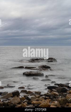 Vertikale Meereslandschaft unter bewölktem Himmel mit Wellen und Felsen im Vordergrund Stockfoto