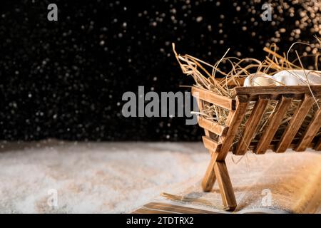 Bibel, Holzkrippe mit Heu und Puppe des Babys auf Schnee vor schwarzem Hintergrund. Das Konzept der Weihnachtsgeschichte Stockfoto