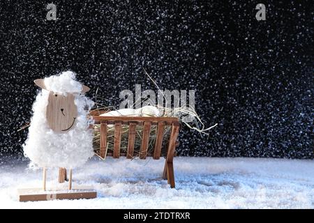 Spielzeug-Schaf und Holzkrippe mit Puppe des Babys auf Schnee vor schwarzem Hintergrund. Das Konzept der Weihnachtsgeschichte Stockfoto