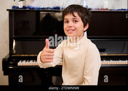 Der hübsche, hübsche kaukasische Teenager sitzt am Flügel und zeigt einen Daumen hoch und blickt in die Kamera. Menschen. Lifestyle. Freizeit und Hobbi Stockfoto