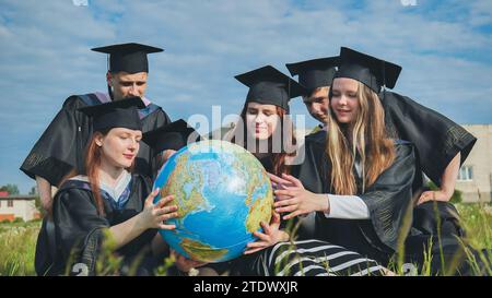 Absolventen in schwarzen Gewändern untersuchen einen geografischen Globus, der auf dem Gras sitzt. Stockfoto