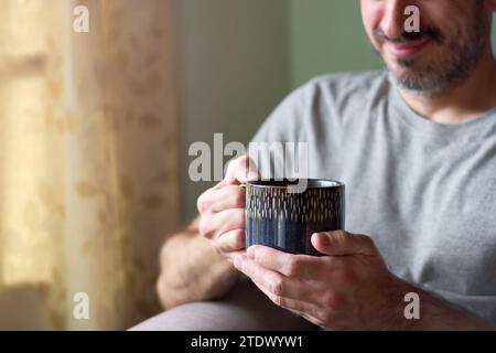 Nahaufnahme eines jungen kaukasischen Mannes, der lächelt und eine Tasse Tee hält. Fröhlicher Mann, der heissen Tee in seinem Wohnzimmer zu Hause genießt und sich während seiner Pause entspannt. C Stockfoto