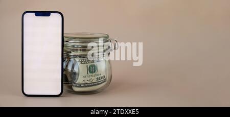 Vertikaler weißer Bildschirm auf modernem Handy im Hintergrund eines Glasgefäßes voller US-Dollar-Banknoten auf beigefarbenem Hintergrund. Bereich für Text. Werbung für Anwendungswebsite. Das Konzept der Geldwirtschaft Banken und Finanzen Stockfoto