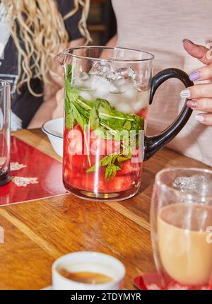 Ein mit Eis gefüllter Krug und ein erfrischendes Wassermelonen-Minzgetränk, das in ein Glas gegossen wird. Die hellen Farben und natürlichen Zutaten sorgen für ein Gefühl von Koh Stockfoto