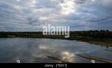 Das Bild bietet einen weiten Blick auf ein ruhiges Gewässer, das einen bewölkten Himmel reflektiert, mit einem herbstlichen Wald im Hintergrund. Die Wasseroberfläche spiegelt die grauen Wolken und blauen Himmelsstreifen wider, während eine Landspieße ins Wasser ragt und den Blick auf die lebendigen Bäume am fernen Ufer lenkt. Die Stille des Wassers steht im Kontrast zum dynamischen Himmel und lässt einen Moment der Ruhe inmitten der wechselnden Jahreszeit vermuten. Die Herbstfarben des Waldes verleihen den ansonsten kühlen Tönen der Szene Wärme. Reflektierende Wasserlandschaft mit herbstlichem Wald und bewölktem Himmel. Hochwertige Fotos Stockfoto