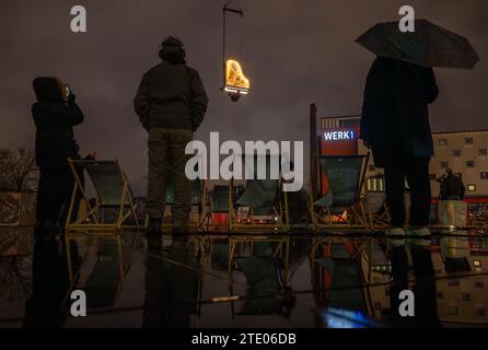 München, Deutschland. Dezember 2023. Zuschauer sehen die Vorschau des Künstlers Alain Roche, der jeden Morgen kurz vor Sonnenaufgang 182 Tage lang im Werksviertel in 10 Metern Höhe ein Klavierkonzert geben will. Der beleuchtete Flügel wird von einem Kran gehalten. Die Konzerte mit dem Titel „wenn die Sonne still steht“ sollen als Förderprojekt für den Bau der Münchner Konzerthalle dienen. Quelle: Peter Kneffel/dpa/Alamy Live News Stockfoto