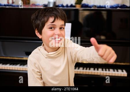 Fröhlicher Schuljunge im Grundalter, der sich hochstellt und lächelnd in die Kamera schaut, während des Musikunterrichts am Klavier sitzt. Süßer und glücklicher, talentierter Junge, A Stockfoto