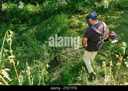Gärtner mähen Unkraut im Garten mit einem Rasentrimmer. Mann mit Rasenmäher im Garten, der grünes Gras schneidet. Stockfoto