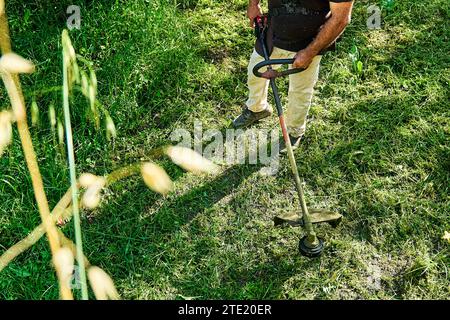 Gärtner mähen Unkraut im Garten mit einem Rasentrimmer. Mann mit Rasenmäher im Garten, der grünes Gras schneidet. Stockfoto