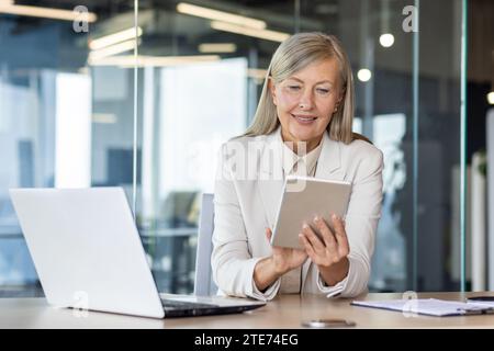 Frau Chefin am Arbeitsplatz im Büro, erfahrene, grauhaarige Geschäftsfrau benutzt Tablet-Computer, prüft gerne Finanzdaten, Bankerin Finanzkraft mit Laptop. Stockfoto