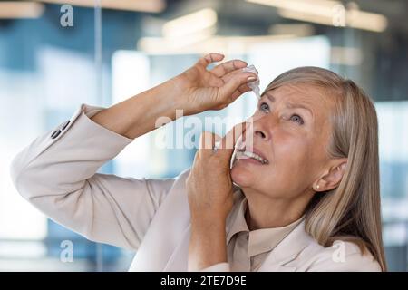 Verängstigte Geschäftsfrau verwendet Augentropfen, grauhaarige Frau übermüdete Wunden Augen reiben Medizin am Arbeitsplatz im Büro, lange Arbeit mit Laptop. Stockfoto