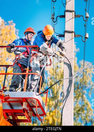 Zwei professionelle Elektriker in Schutzhelmen reparieren Stromleitungen von der Ladestation des Schaufelwagens. Ansicht von unten. Elektriker wechseln Kabel an Straßenbeleuchtungsmasten. Stockfoto