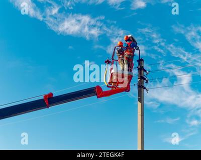 Zwei Elektriker von der Halterung der Antennenplattform oder des Krans reparieren die Straßenbeleuchtung. Professionelle Elektriker tragen Helme, Overalls und Versicherungsarbeiten in der Höhe. Ansicht der Arbeiter von unten nach oben zum Himmel. Authentischer Workflow. Stockfoto
