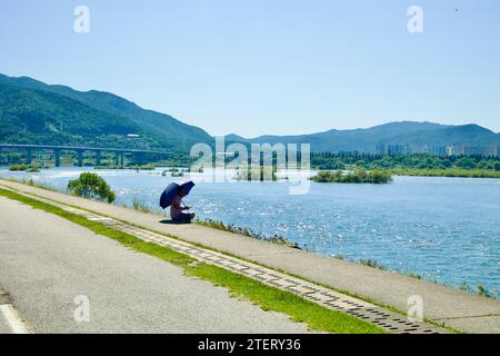 Namyangju City, Südkorea - 30. September 2023: Eine Einzelfigur mit einem Regenschirm sitzt am Han River und betrachtet den ruhigen Blick auf den kleinen isl Stockfoto