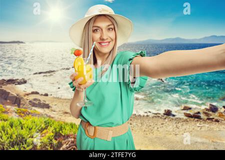 Schöne junge Frau in grünem Kleid, die einen Cocktail hält und ein Selfie am Strand in Griechenland macht Stockfoto